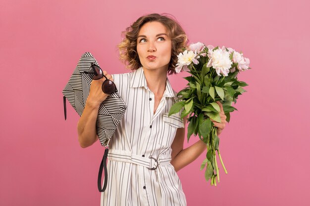 Attraktive stilvolle Frau im eleganten weißen gestreiften Kleid auf rosa Studiohintergrund emotionaler Gesichtsausdruck, überrascht, Handtasche, Blumenstrauß, lustige, lockige Frisur, Modesommer-Trendzubehör