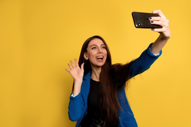 Attraktive stilvolle Frau mit dunklem Haar, das blaue Jacke trägt, die winkt und Selfie macht. Freudiges Mädchen mit hellbraunem Haar, das Selfie mit sanftem Lächeln auf gelber Wand macht