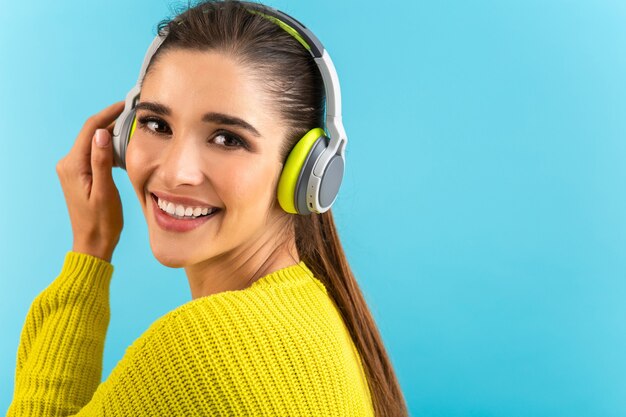 Attraktive stilvolle junge Frau, die Musik in den kabellosen Kopfhörern hört, die glücklich tragen, gelben Strickpullover bunte Artmode, die lokal auf blauem Hintergrund aufwirft