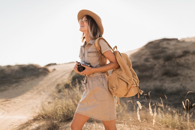 Attraktive stilvolle junge Frau im Khaki-Kleid in der Wüste, die in Afrika auf Safari reist, Hut und Rucksack trägt und Foto auf Vintage-Kamera macht