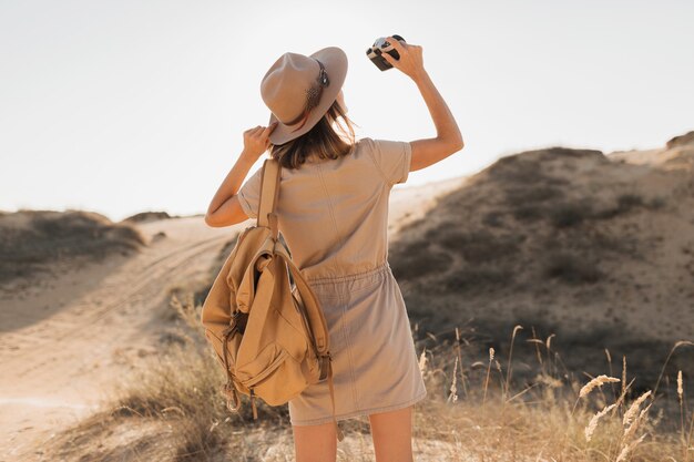 Attraktive stilvolle junge Frau im Khaki-Kleid in der Wüste, die in Afrika auf Safari reist, Hut und Rucksack trägt und Foto auf Vintage-Kamera macht