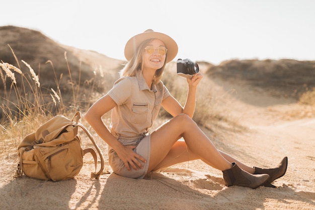 Attraktive stilvolle junge Frau im Khaki-Kleid in der Wüste, die in Afrika auf Safari reist, Hut und Rucksack trägt und Foto auf Vintage-Kamera macht