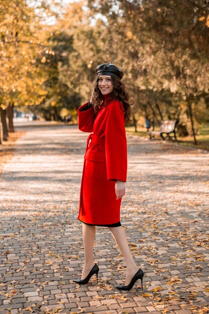 Attraktive stilvolle lächelnde dünne Frau mit dem lockigen Haar, das im Park geht, gekleidet im warmen roten Mantel herbst trendige Mode, Straßenart, Baskenmützenhut tragend