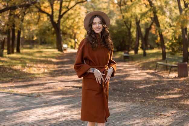 Attraktive stilvolle lächelnde dünne Frau mit dem lockigen Haar, das im Park geht, gekleidet in warmem braunem Mantel, herbsttrendiger modischer Straßenstil