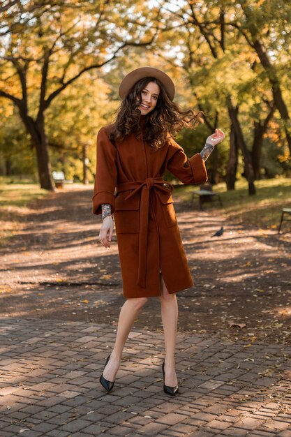 Attraktive stilvolle lächelnde dünne Frau mit dem lockigen Haar, das im Park geht, gekleidet in warmem braunem Mantel, herbsttrendiger modischer Straßenstil