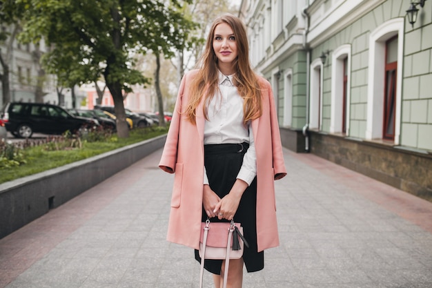 Attraktive stilvolle lächelnde Frau, die Stadtstraße im rosa Mantel geht