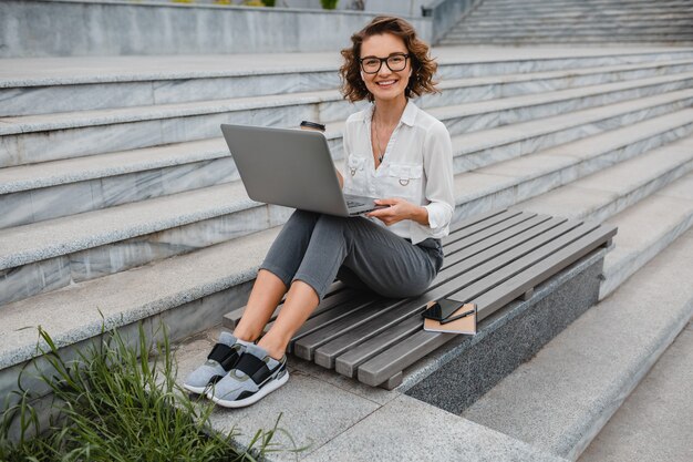 Attraktive stilvolle lächelnde Frau mit Brille, die am Laptop schreibt