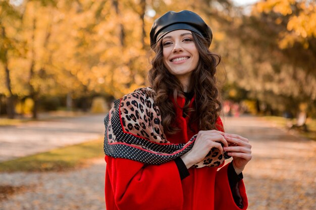 Attraktive stilvolle lächelnde Frau mit dem lockigen Haar, das im Park geht, gekleidet im warmen roten Mantel herbst trendige Mode, Straßenart, tragenden Baskenmützenhut und Schal mit Leopardenmuster