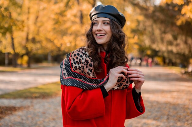 Attraktive stilvolle lächelnde Frau mit dem lockigen Haar, das im Park geht, gekleidet im warmen roten Mantel herbst trendige Mode, Straßenart, tragenden Baskenmützenhut und Schal mit Leopardenmuster