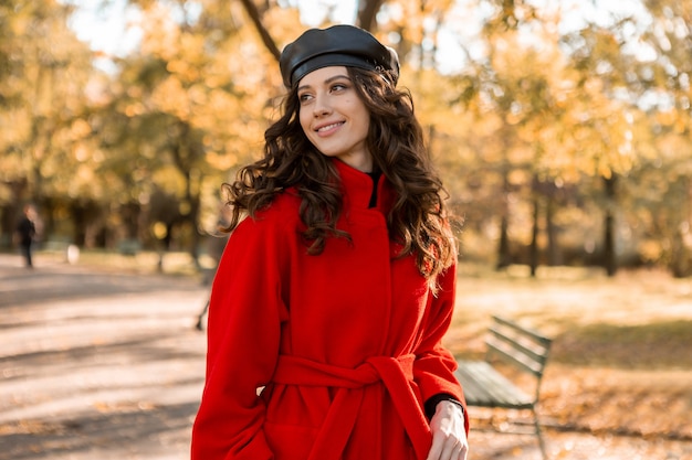 Attraktive stilvolle lächelnde Frau mit dem lockigen Haar, das im Park geht, gekleidet im warmen roten Mantel herbstliche modische Mode, Straßenart, Baskenmützenhut tragend