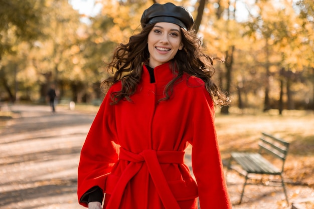 Attraktive stilvolle lächelnde Frau mit dem lockigen Haar, das im Park geht, gekleidet im warmen roten Mantel herbstliche modische Mode, Straßenart, Baskenmützenhut tragend