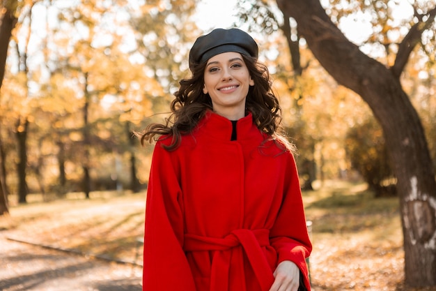 Attraktive stilvolle lächelnde Frau mit dem lockigen Haar, das im Park geht, gekleidet im warmen roten Mantel herbstliche modische Mode, Straßenart, Baskenmützenhut tragend