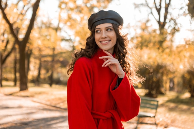 Attraktive stilvolle lächelnde Frau mit dem lockigen Haar, das im Park geht, gekleidet im warmen roten Mantel herbstliche modische Mode, Straßenart, Baskenmützenhut tragend