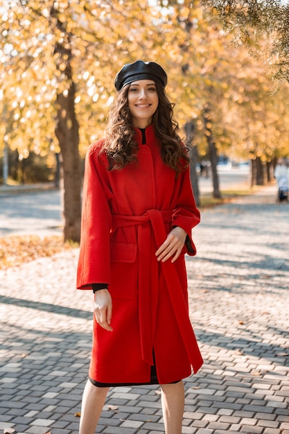 Attraktive stilvolle lächelnde Frau mit dem lockigen Haar, das im Park geht, gekleidet im warmen roten Mantel herbstliche modische Mode, Straßenart, Baskenmützenhut tragend