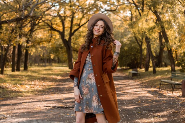 Attraktive stilvolle lächelnde Frau mit dem lockigen Haar, das im Park geht, gekleidet in bedrucktes Kleid und warme Mantelherbsttrendmode, Straßenart