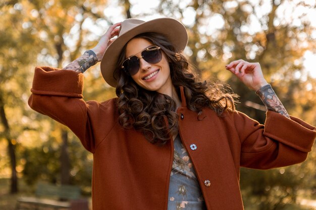 Attraktive stilvolle lächelnde Frau mit dem lockigen Haar, das im Straßenpark geht, gekleidet in warmer brauner Mantelherbsttrendmode, Straßenart, die Hut und Sonnenbrille trägt