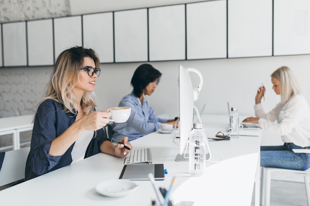 Attraktive Studentin mit blonden Haaren, die sich auf Prüfungen vorbereiten, in der Nähe des Computers sitzen und Latte trinken