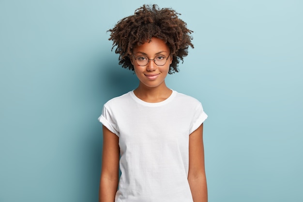 Attraktive Studentin mit lockigem Haar, trägt transparente Brille, weißes T-Shirt, steht vor blauem Hintergrund, hat ruhigen Gesichtsausdruck, zartes Lächeln,