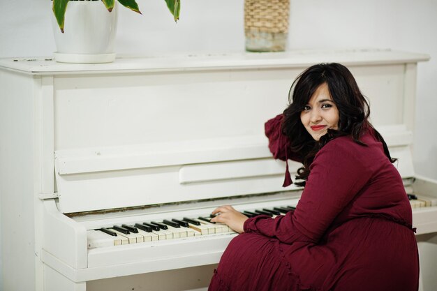 Attraktive südasiatische Frau in tiefrotem Kleid posiert im Zimmer und spielt auf dem Klavier