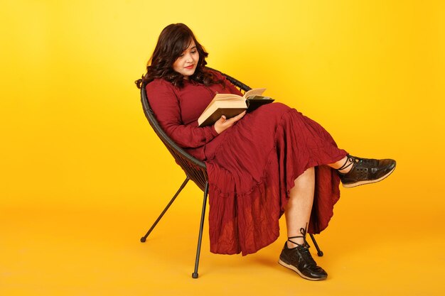 Attraktive südasiatische Frau in tiefrotem Kleid posierte im Studio auf gelbem Hintergrund, sitzend auf Stuhl mit Buch
