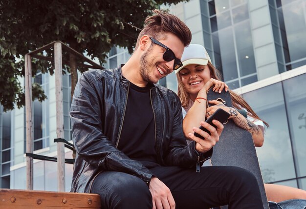 Attraktive, trendig gekleidete junge Paare haben Spaß zusammen, während sie zusammen auf einer Bank in der Nähe des Wolkenkratzers sitzen.