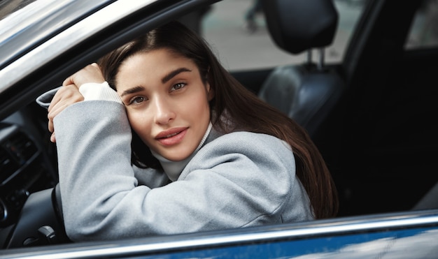 Attraktive und elegante Fahrerin, die sich in ihrem Auto ausruht, sich auf das Autorad stützt und aus dem Fenster schaut und auf dem Parkplatz wartet