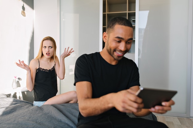 Attraktive verärgerte lustige junge Frau auf dem Bett ist wütend auf aufgeregten glücklichen Kerl, der mit Tablette auf Bett zu Hause in der modernen Wohnung spielt. Beziehung, beleidigtes Mädchen, Spaß haben, Unverständnis