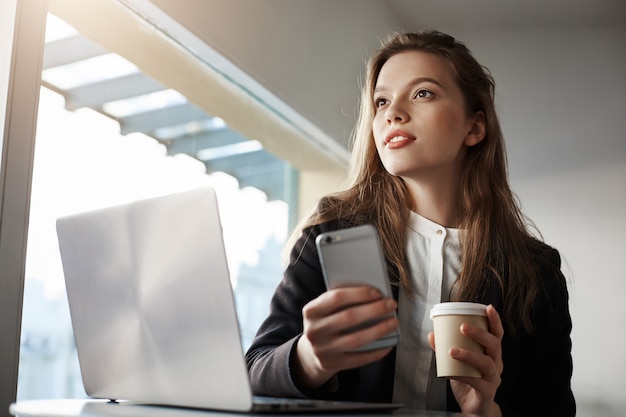 attraktive weibliche Büroangestellte, die im Café mit Laptop sitzt, Kaffee trinkt und Smartphone hält, träumt, während sie beiseite schaut