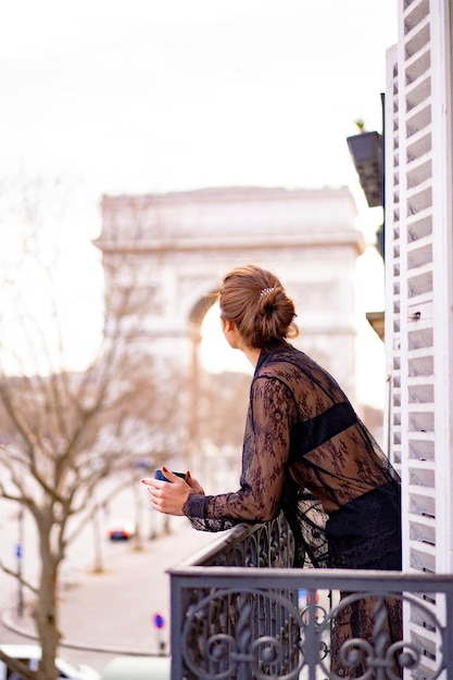 Attraktive Yang-Frau im Schlafanzug trinkt morgens Kaffee auf dem Balkon in der Stadt Paris. Blick auf den Triumphbogen.