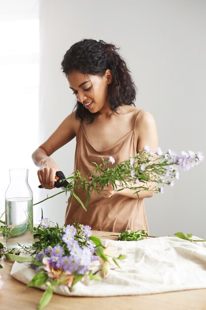 Attraktiver afrikanischer weiblicher Florist, der Stämme am Arbeitsplatz schneidet. Weiße Wand.