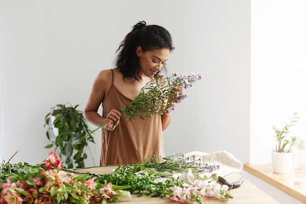 Attraktiver afrikanischer weiblicher Florist lächelnd, der Blumenstrauß am Arbeitsplatz über weißer Wand macht.