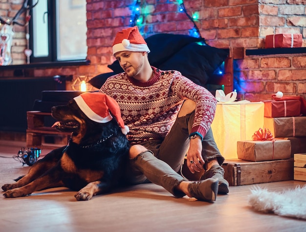 Attraktiver bärtiger Hipster-Mann sitzt mit seinem Rottweiler-Hund auf einem Boden in einem Raum mit Weihnachtsdekoration.