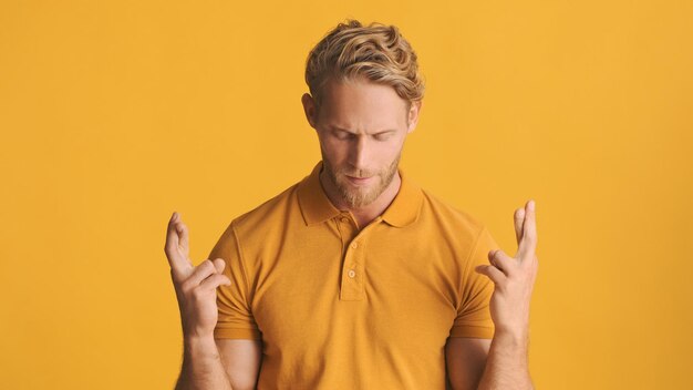 Attraktiver blonder bärtiger Mann mit gekreuzten Fingern, der hoffentlich vor der Kamera betet, isoliert auf gelbem Hintergrund