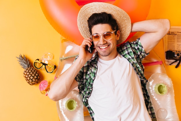 Attraktiver brünetter Mann im karierten Hemd und im weißen T-Shirt mit dem Lächeln, das am Telefon spricht. Mann mit Hut und Sonnenbrille liegt auf aufblasbarer Matratze.