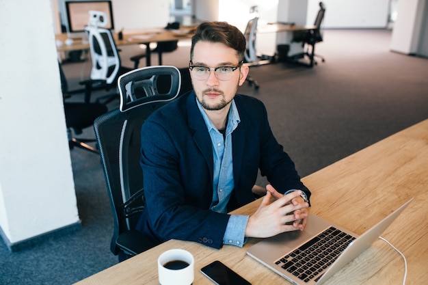 Attraktiver dunkelhaariger Mann sitzt am Tisch im Büro. Er trägt ein blaues Hemd mit einer schwarzen Jacke. Er schaut zur Seite. Sicht von oben.