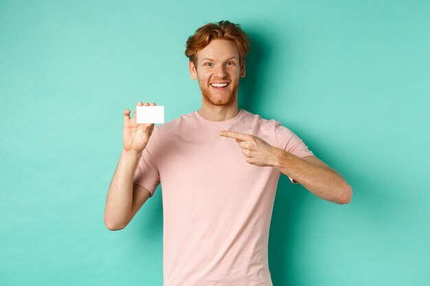 Attraktiver erwachsener Mann mit Bart und roten Haaren, der mit dem Finger auf die Plastikkreditkarte zeigt, erfreut in die Kamera lächelt und über türkisfarbenem Hintergrund steht