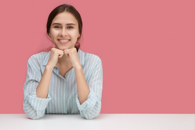 Attraktiver fröhlicher Büroangestellter der jungen Frau mit breitem glücklichem Lächeln, das den Arbeitstag genießt, der am weißen Schreibtisch gegen leere rosa Wand mit Copyspace für Ihren Werbeinhalt sitzt