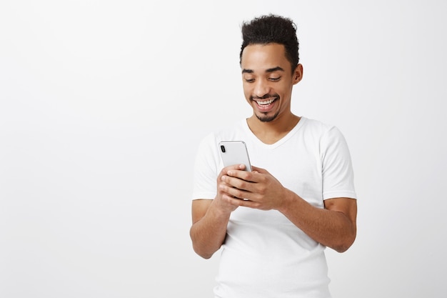Attraktiver glücklicher junger Afroamerikanermann, der Handy verwendet und auf Bildschirm lächelt, Chat, mit Anwendung