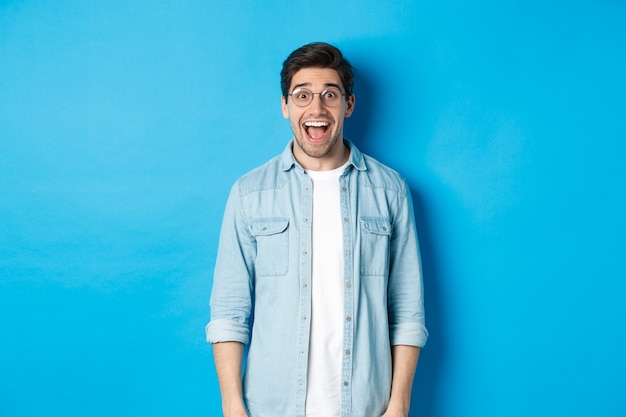 Attraktiver glücklicher Mann mit Brille, der überrascht aussieht, die Werbung überprüft und auf blauem Hintergrund steht.
