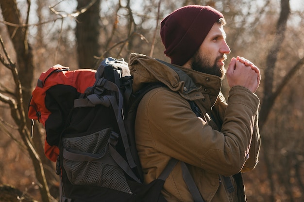 Attraktiver Hipster-Mann, der mit Rucksack im Herbstwald mit warmer Jacke und Hut reist, aktiver Tourist, Natur in der kalten Jahreszeit erforschend