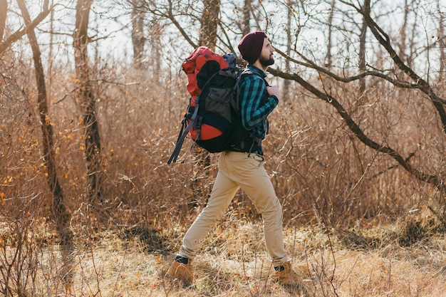 Attraktiver Hipster-Mann, der mit Rucksack im Herbstwald reist und kariertes Hemd und Hut trägt