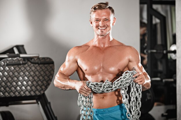 Attraktiver hunky männlicher Bodybuilder, der Bodybuilding-Pose im Fitnessstudio mit Eisenketten tut