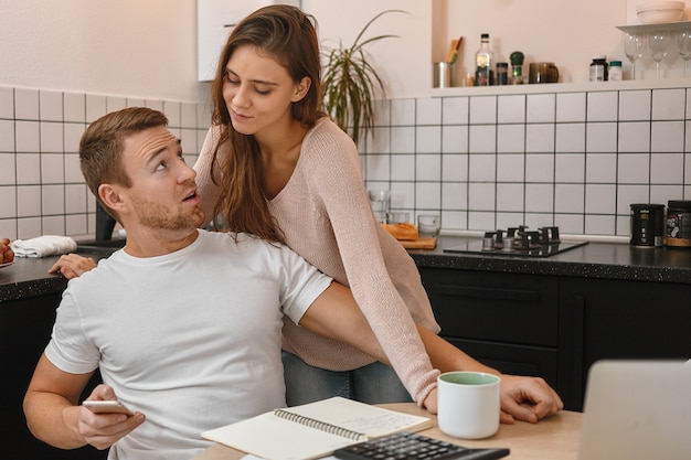 Attraktiver junger bärtiger Mann im weißen T-Shirt, der in der Küche am Tisch mit Papieren, Laptop und Taschenrechner sitzt, Smartphone hält und sich weigert, seiner verdächtigen Frau eine SMS zu schicken. Menschen und Technik