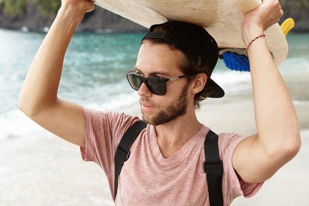 Attraktiver junger bärtiger Surfer in den Schatten, die am Strand am Meer stehen