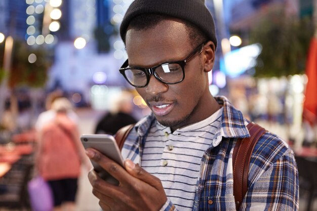 Attraktiver junger dunkelhäutiger europäischer Anhalter mit Rucksack, der eine trendige Brille und einen Hut trägt und Routen und Orte auf der Online-Karte auf seinem Smartphone studiert und mitten in einer fremden Stadt steht
