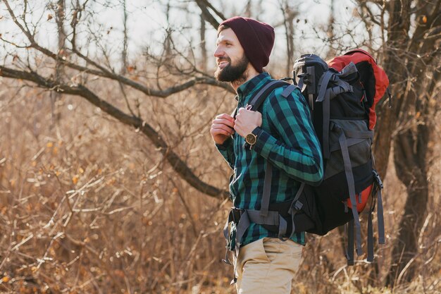 Attraktiver junger Hipster-Mann, der mit Rucksack im Herbstwald reist und kariertes Hemd und Hut trägt