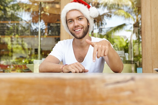 Attraktiver junger kaukasischer bärtiger Mann im weißen T-Shirt und im roten Weihnachtsmannhut, der am Tisch sitzt und gestikuliert, seinen Zeigefinger nach unten zeigt und einen glücklichen und fröhlichen Ausdruck auf seinem Gesicht hat