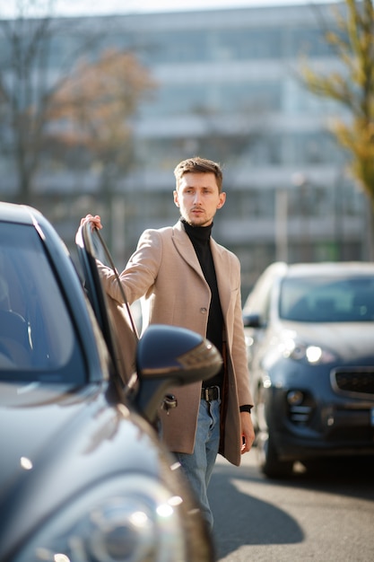 Attraktiver junger kaukasischer Mann öffnet eine Autotür, gekleidet in beigem Mantel in der Innenstadt an einem sonnigen Tag