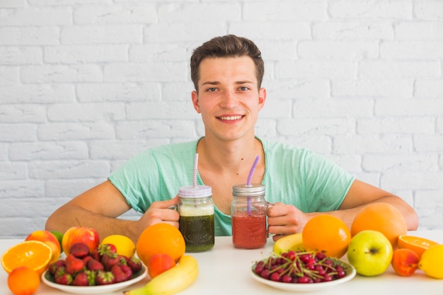 Attraktiver junger Mann, der grüne und rote Smoothiegläser mit bunten Früchten auf Tabelle hält