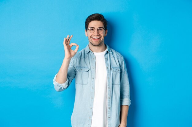 Attraktiver junger Mann mit Brille und Freizeitkleidung, der ein gutes Zeichen in Genehmigung zeigt, wie etwas, das vor blauem Hintergrund steht